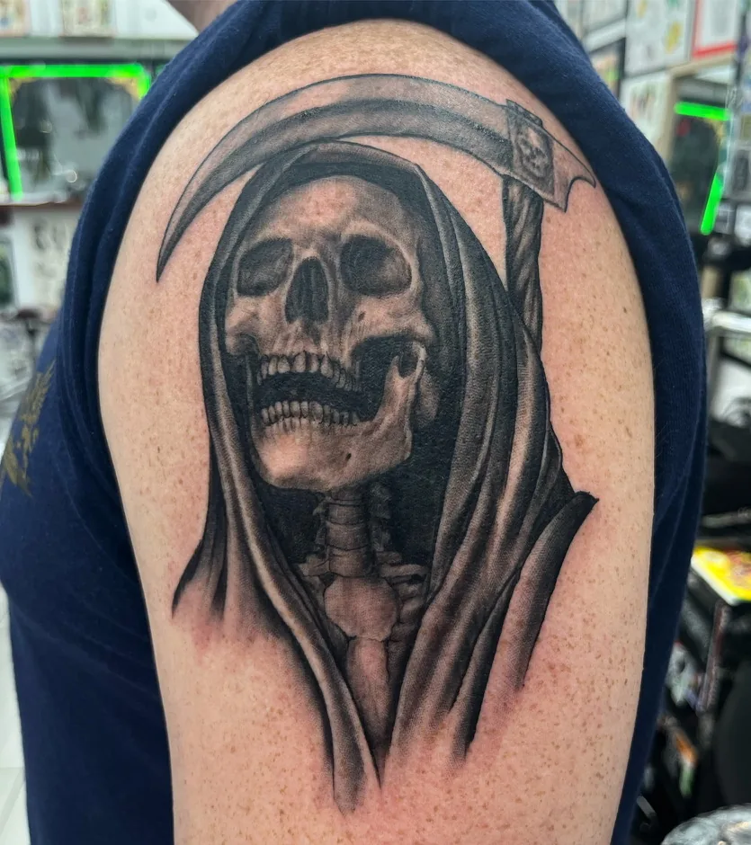 Realism tattoo by Heather Baldwin — cloaked grim reaper skull holding a scythe with a small carved skull at the blade, black and grey realism on the shoulder