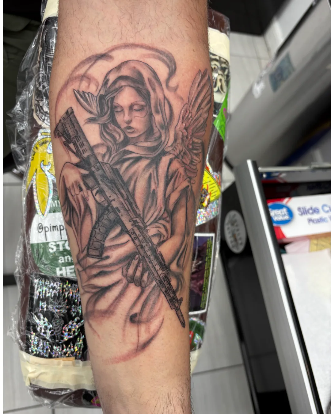 Tattoo by Heather Baldwin — hooded guardian angel holding a rifle, soft grey shading on the forearm, military memorial style