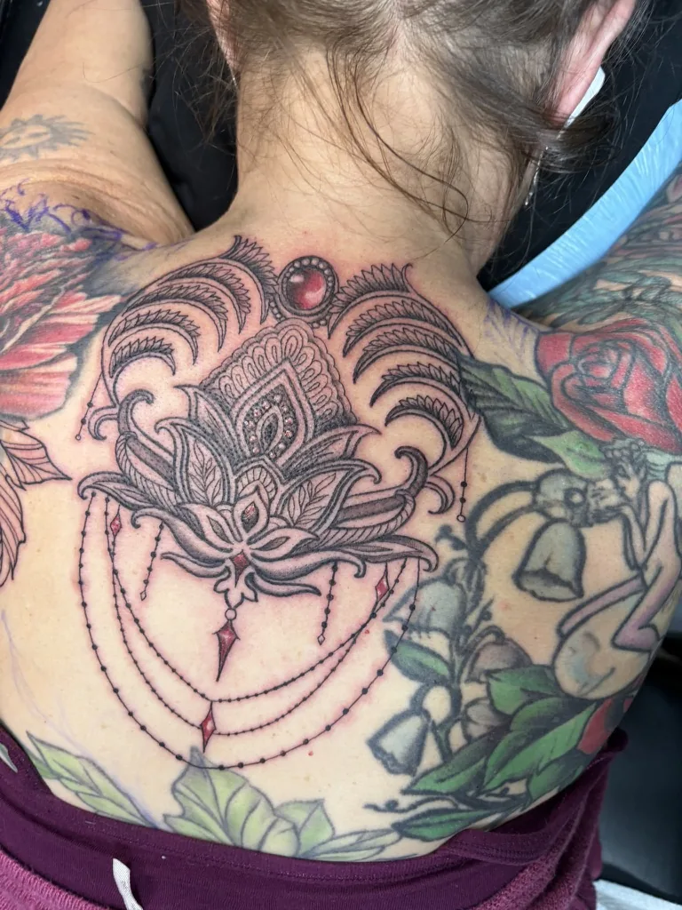 Ornamental tattoo by Heather Baldwin — lotus and ruby medallion with draped jewelry chains across the upper back and base of the neck, color fine-line on existing skin