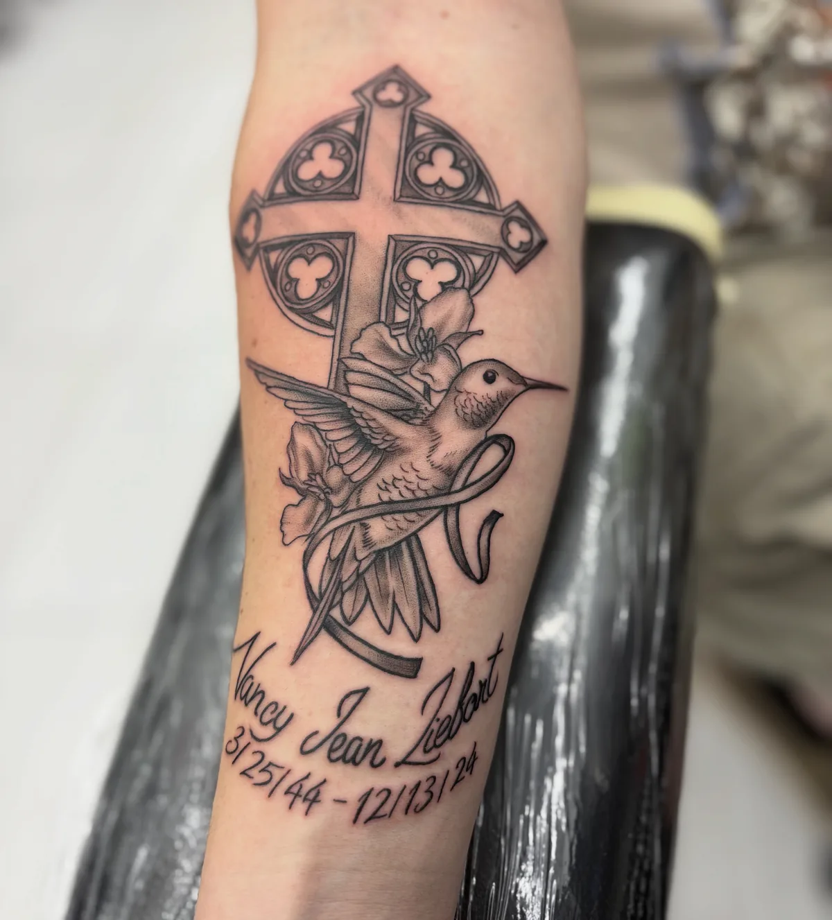 Memorial tattoo by Heather Baldwin — Celtic cross above a hummingbird and lily, name banner 'Nancy Jean Liebert' below, fine-line black and grey on the forearm