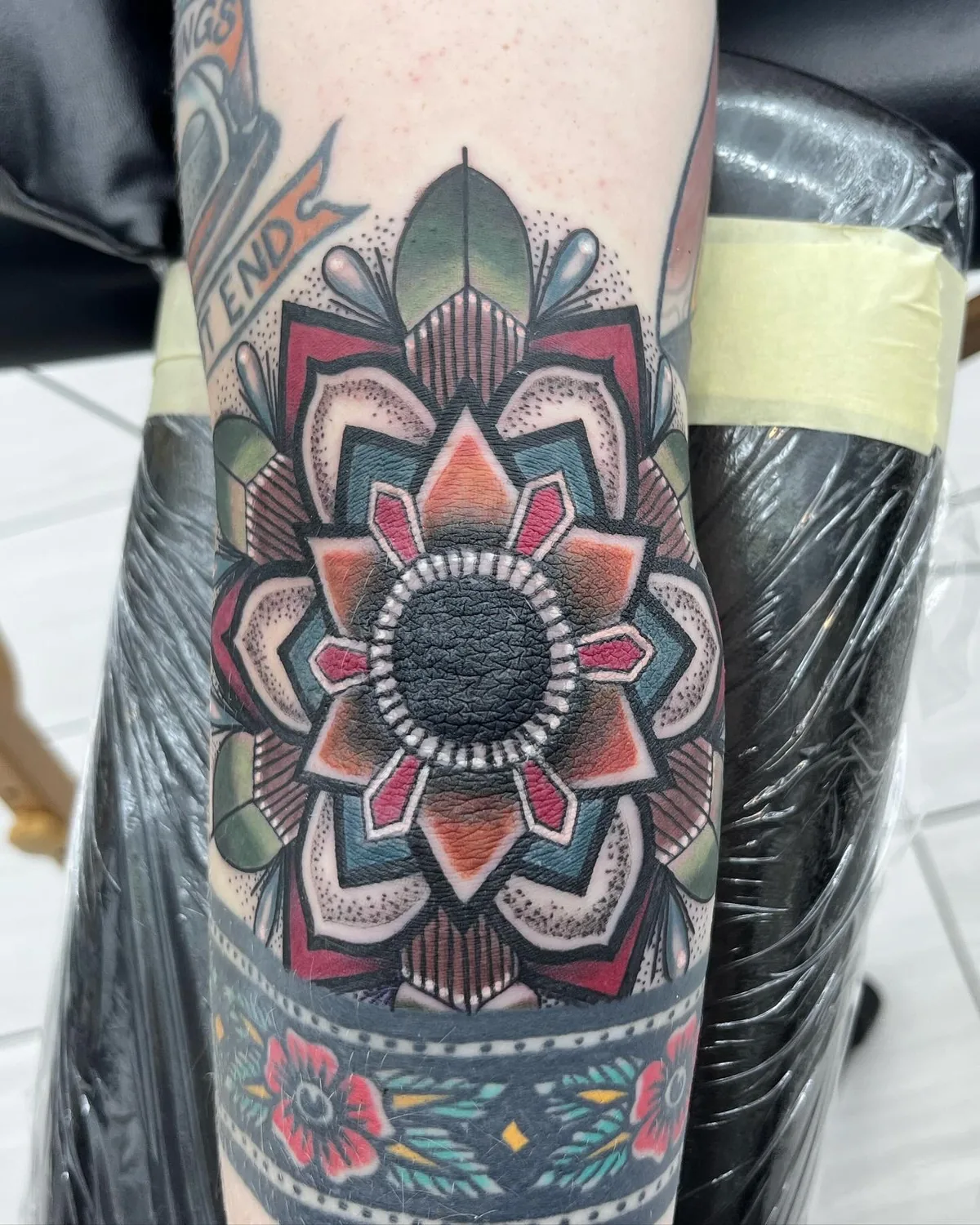 Neo-traditional tattoo by Heather Baldwin — bold color mandala medallion centered over the elbow with dotwork shading and geometric petals