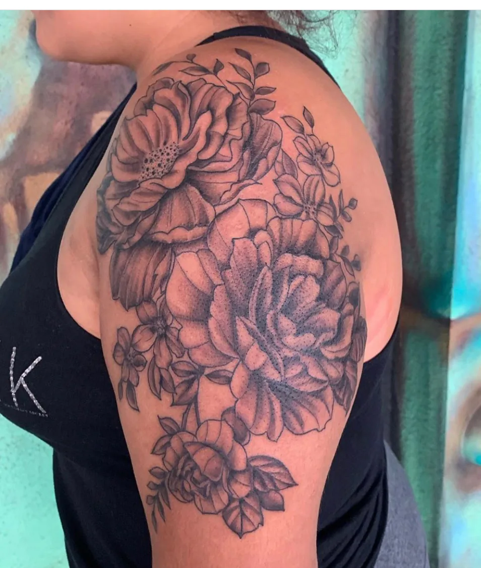 Floral tattoo by Heather Baldwin — full peony and rose bouquet wrapping the shoulder and upper arm in detailed black and grey