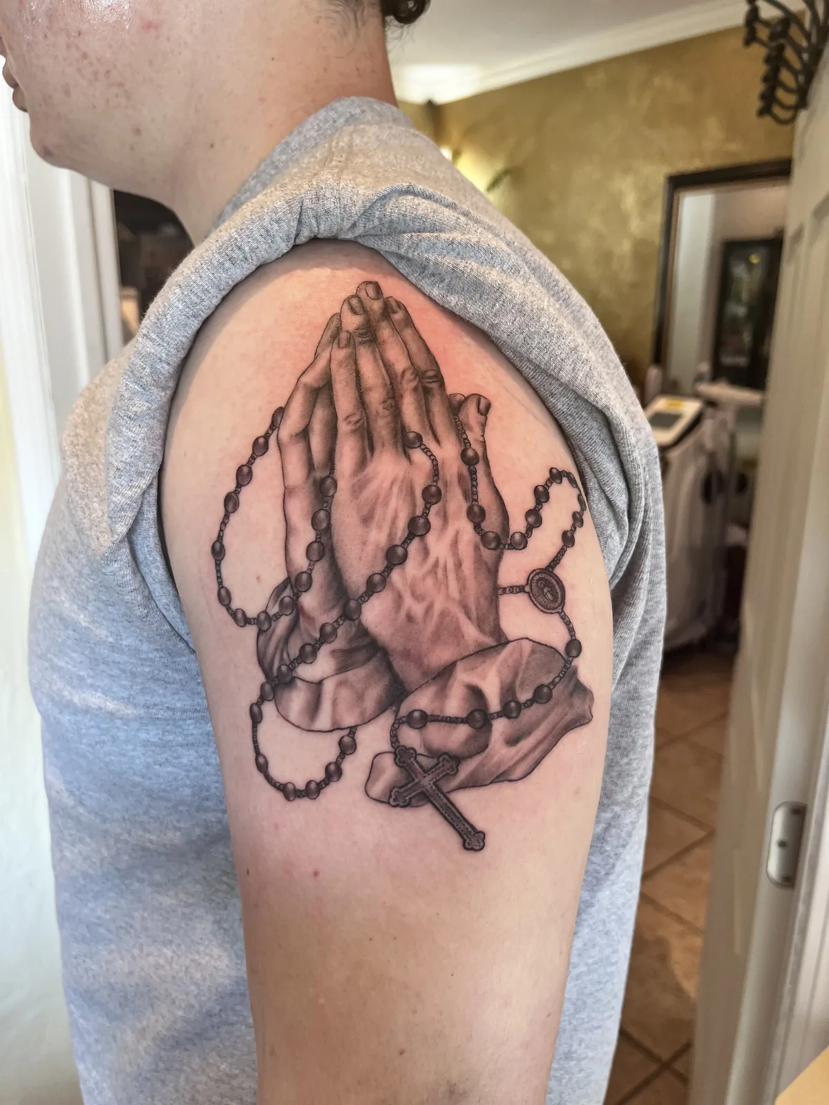 Praying hands tattoo by Heather Baldwin — clasped hands wrapped in a rosary, crucifix below, B&G realism on the bicep