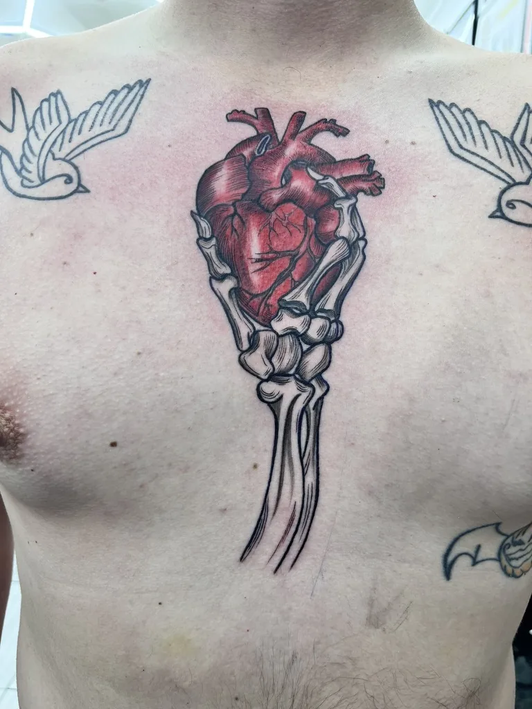 Illustrative tattoo by Heather Baldwin — skeletal hand clutching an anatomical heart between two outlined sparrows on the chest, color realism on the heart
