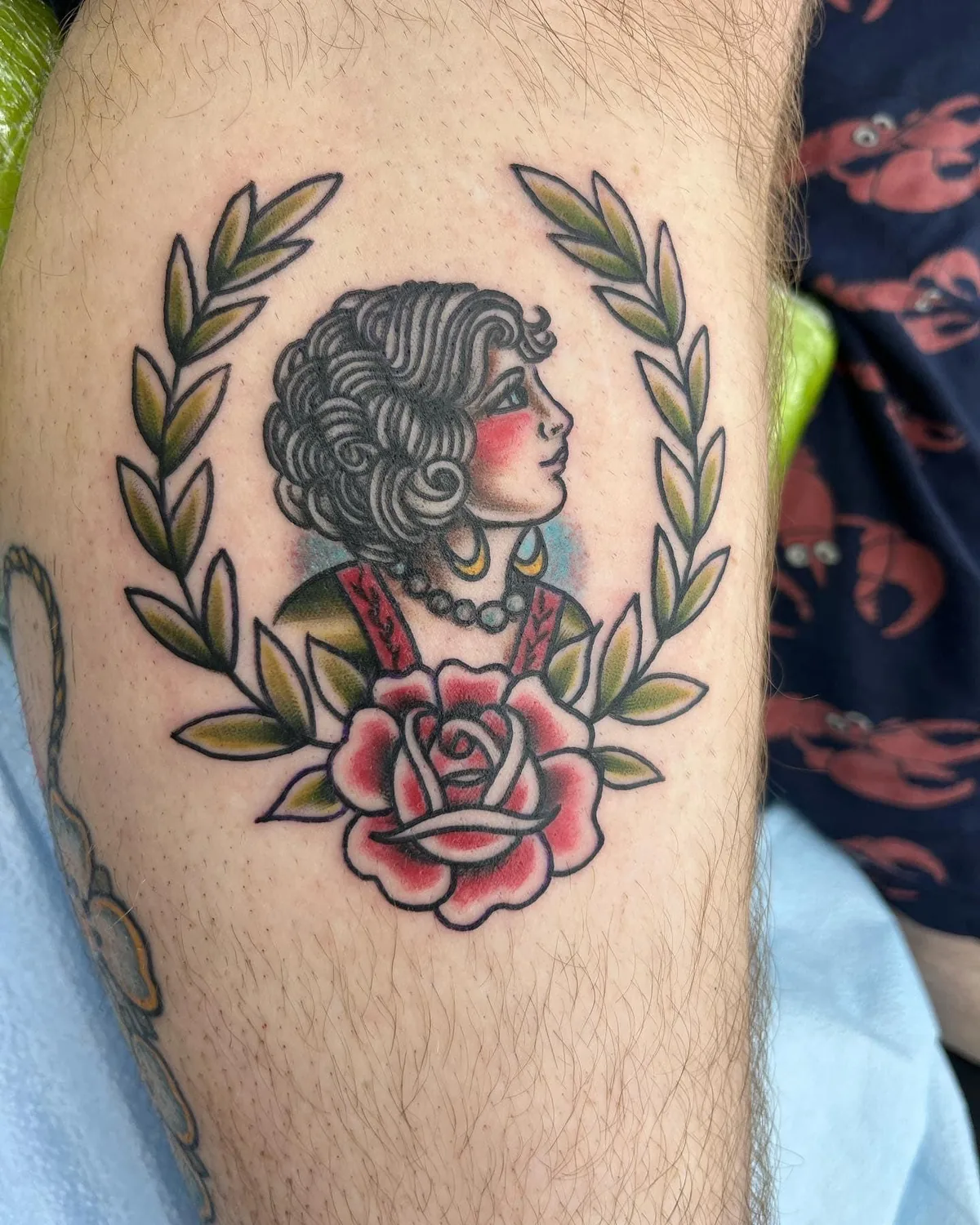 American Traditional tattoo by Heather Baldwin — classic girl head with curls and earrings framed by a laurel wreath and a single rose, color trad on the thigh