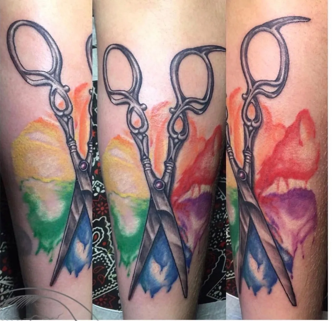 Watercolor tattoo by Heather Baldwin — open hairdresser shears with a rainbow watercolor wash behind them, color illustrative on the forearm
